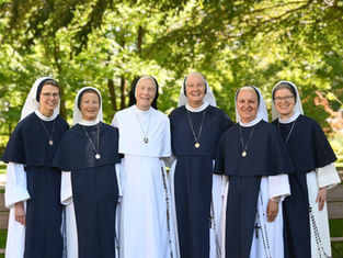 Six nuns in navy and white habits smile together outdoors on a sunny day, with green trees in the background, exuding a joyful mood.