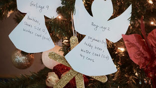 Paper angel ornaments with gift wishes for children hang on a decorated Christmas tree. Lights and red flowers in the background.
