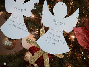 Paper angel ornaments with gift wishes for children hang on a decorated Christmas tree. Lights and red flowers in the background.