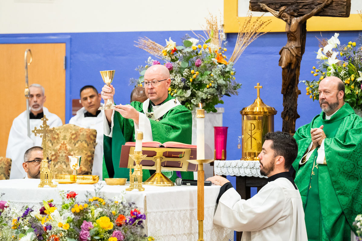 Bishop in green vestments lifts a chalice at a Catholic altar with candles, flowers, crucifix, and seated clergy.