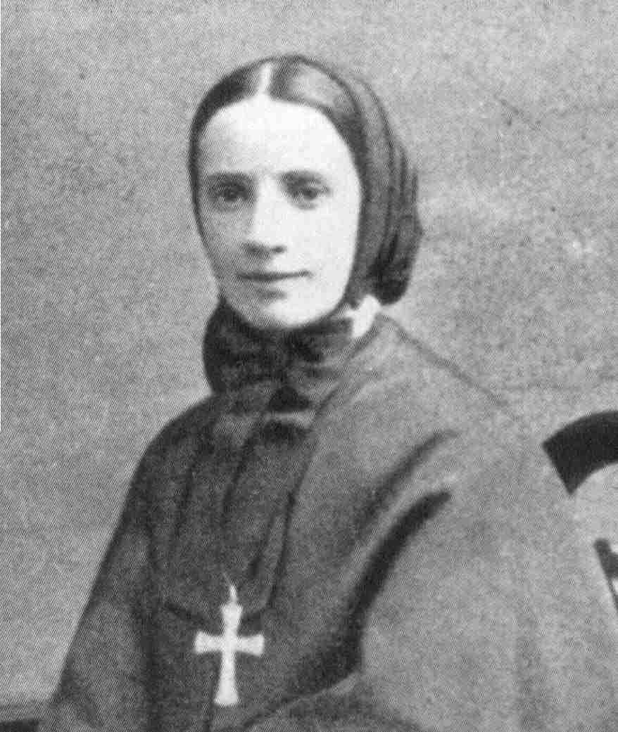 Black-and-white portrait of a woman in a headscarf and dark dress, wearing a cross pendant, against a plain background.
