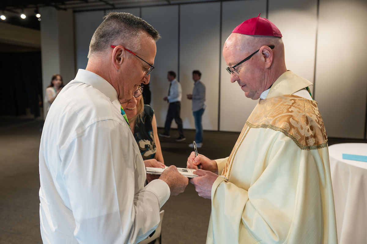 A man in religious attire signs a book for another man in a white shirt. Neutral indoor setting with people blurred in the background.