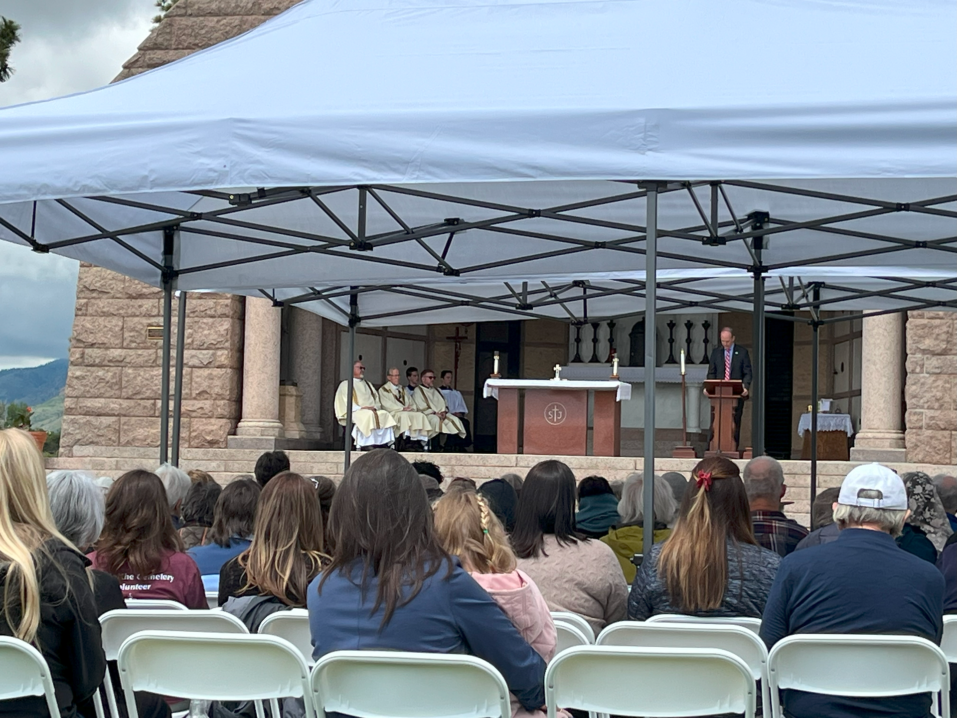 Outdoor ceremony with people seated under a tent, a speaker at a podium, and religious figures in robes on a stage. Stone building backdrop.