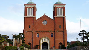 Red brick church with two towers and white arched windows, set against a blue sky. Lush greenery on either side of the entrance.