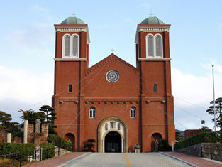 Red brick church with two towers and white arched windows, set against a blue sky. Lush greenery on either side of the entrance.