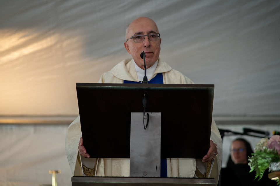 A priest in white vestments speaks from a lectern during a church gathering inside a tent.