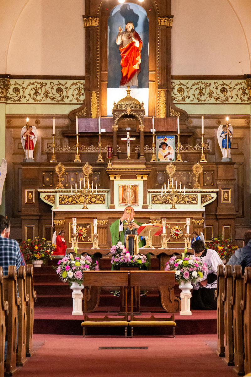 Priest in green robes holds Eucharist at ornate church altar with candles, flowers, and religious figurines. Congregation sits in pews.