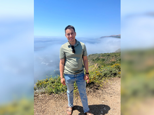 A person smiling in a green shirt stands on a rocky path with foggy mountains and blue sky in the background, conveying a serene mood.
