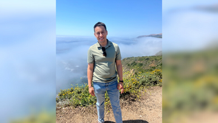A person smiling in a green shirt stands on a rocky path with foggy mountains and blue sky in the background, conveying a serene mood.