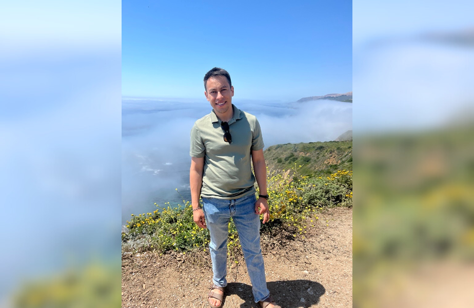 A person smiling in a green shirt stands on a rocky path with foggy mountains and blue sky in the background, conveying a serene mood.