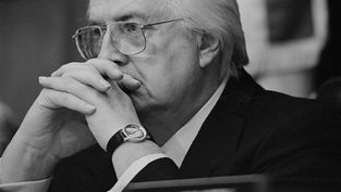 Elderly man in suit and glasses contemplates with hands clasped. Wears watch, sitting in a blurred indoor setting, serious mood.
