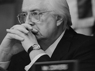 Elderly man in suit and glasses contemplates with hands clasped. Wears watch, sitting in a blurred indoor setting, serious mood.