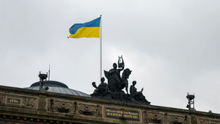 Ukrainian flag waves above historic building adorned with sculptures under a gray sky. Visible inscriptions: "1748" and "1874". Mood: solemn.