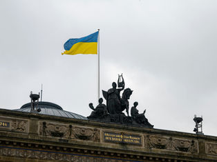 Ukrainian flag waves above historic building adorned with sculptures under a gray sky. Visible inscriptions: "1748" and "1874". Mood: solemn.