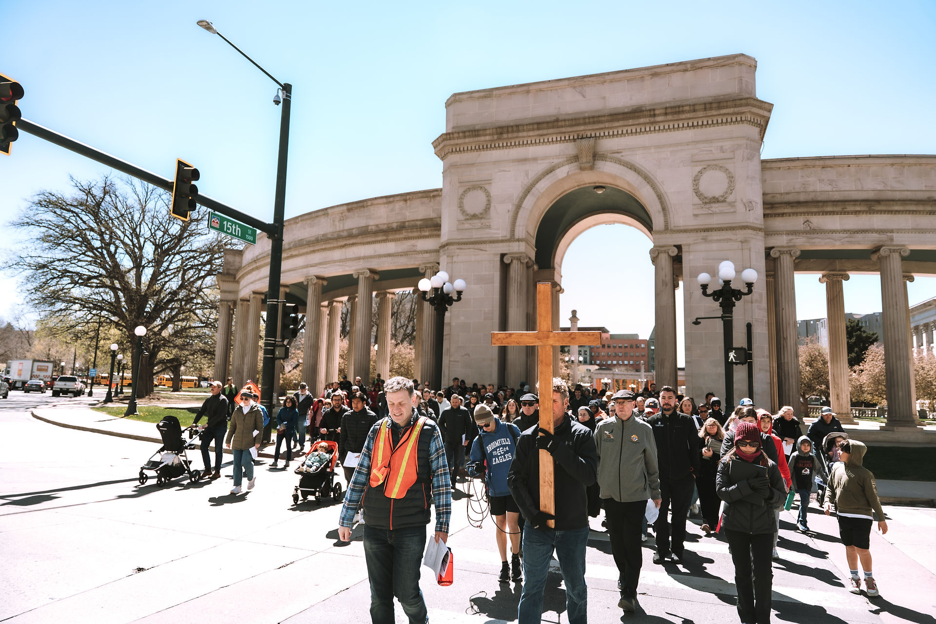 A large group walks past a civic colonnade, following a wooden cross during a public procession.