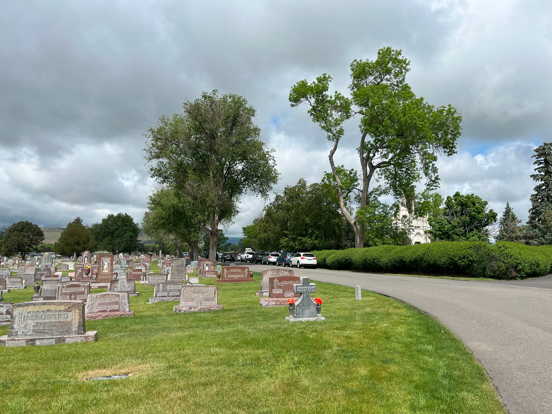 A cemetery road stretches into the distance, with headstones and well-trimmed grass visible on the side of the path. It's a cloudy day.