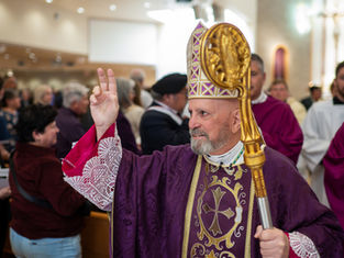 Bishop in ornate purple robe and mitre holds up two fingers in blessing. He carries a golden staff. People gather in a church setting.