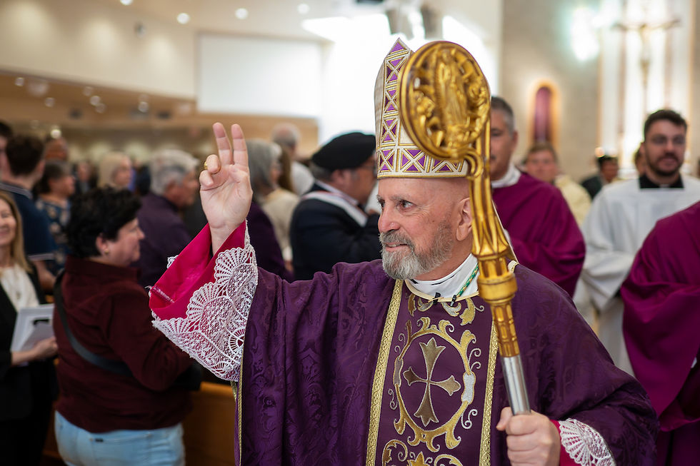 Bishop in ornate purple robe and mitre holds up two fingers in blessing. He carries a golden staff. People gather in a church setting.