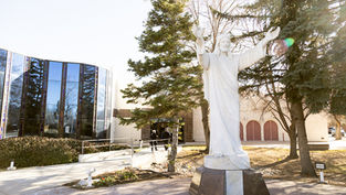White statue of Jesus with arms raised in front of a modern glass building, surrounded by trees. Bright, sunny day.
