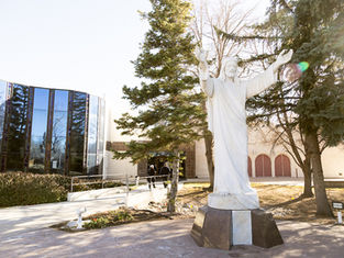 White statue of Jesus with arms raised in front of a modern glass building, surrounded by trees. Bright, sunny day.
