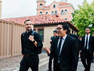 Group of men in suits and clerical attire walking and chatting outside, with brick building and greenery in the background. Casual mood.