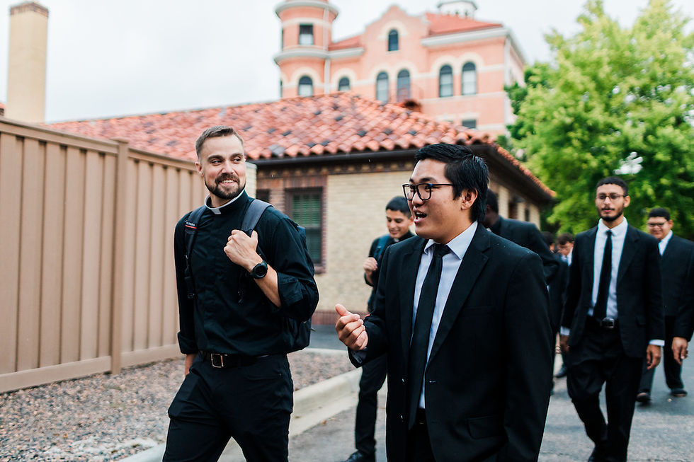 Group of men in suits and clerical attire walking and chatting outside, with brick building and greenery in the background. Casual mood.