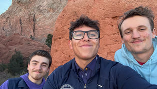 Three smiling people take a selfie against a red rock formation. One wears glasses and a jacket; another a blue hoodie. Clear sky background.