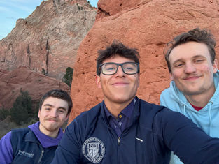 Three smiling people take a selfie against a red rock formation. One wears glasses and a jacket; another a blue hoodie. Clear sky background.