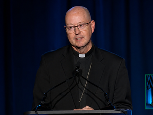 Bald bishop in glasses speaks at podium on blue-lit stage, with sign-language interpreter inset at lower right.