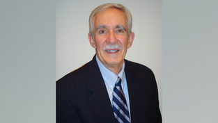 Man with gray hair and mustache smiling, wearing a dark suit and striped tie. Plain light gray background, conveying a professional mood.