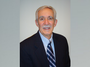 Man with gray hair and mustache smiling, wearing a dark suit and striped tie. Plain light gray background, conveying a professional mood.