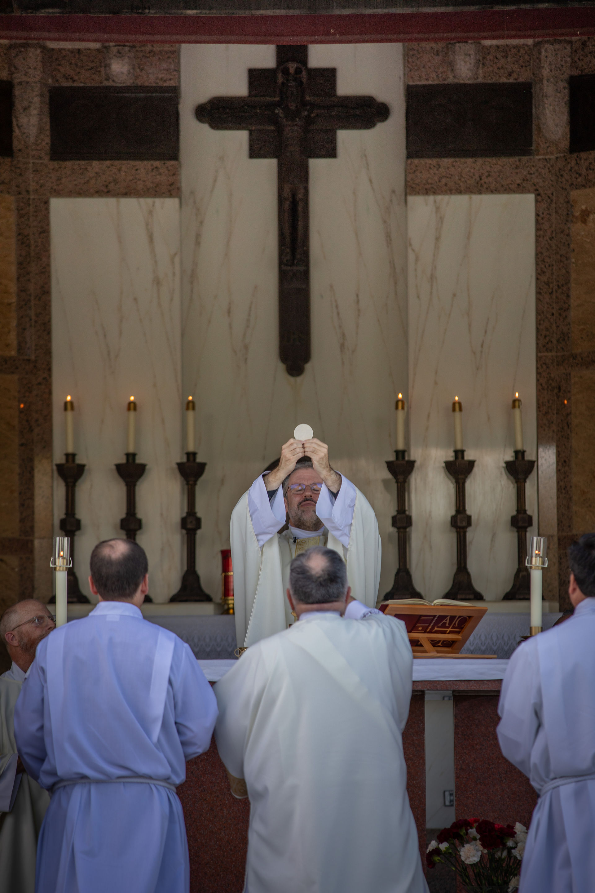 Priest raises Eucharist during Mass; four others look on. Altar with candles and cross in background. Serene, reverent atmosphere.