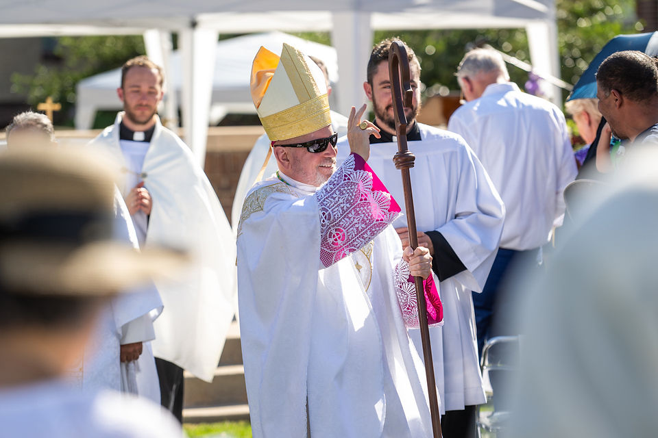 Bishop in white vestments and gold mitre smiles, holding a staff and blessing outdoors amid other clergy and onlookers.