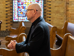A person in a black clerical outfit prays in a church pew, with colorful stained glass in the background. The setting is solemn and introspective.