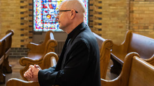 A person in a black clerical outfit prays in a church pew, with colorful stained glass in the background. The setting is solemn and introspective.