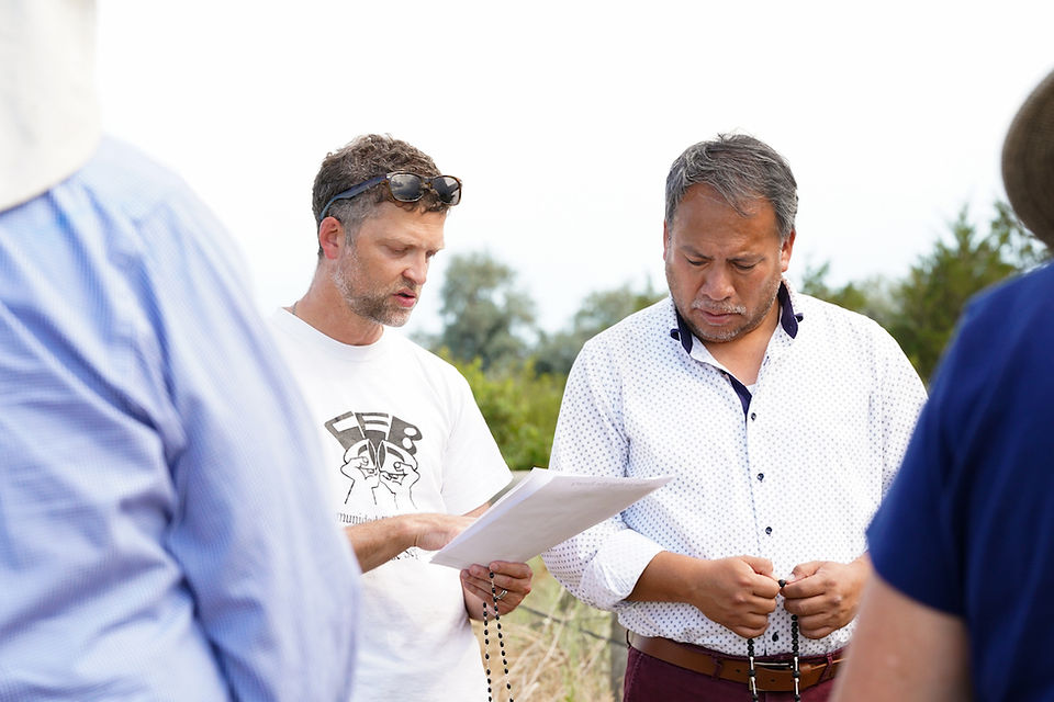 Two men outdoors study a paper, one holding rosary beads, with blurred people around them under a bright sky.