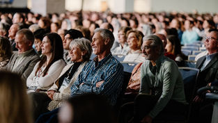 Audience seated in a large hall, smiling and attentive. Rows of people create a sense of community and interest. Neutral background.