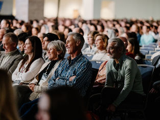 Audience seated in a large hall, smiling and attentive. Rows of people create a sense of community and interest. Neutral background.