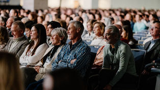 Audience seated in a large hall, smiling and attentive. Rows of people create a sense of community and interest. Neutral background.