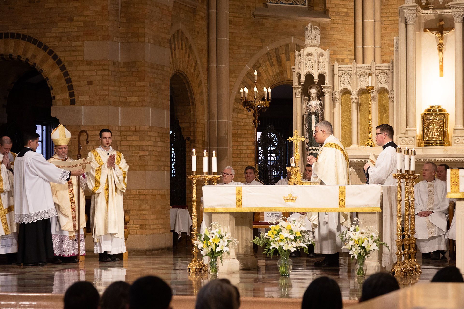 A priest converses with an archbishop during a religious ceremony. The background features the beautiful, ornate chapel complete with tan brickwork. The mood is celebratory and excited.