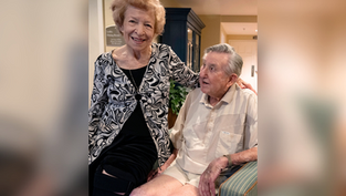 Elderly woman in patterned blouse smiles, stands beside seated man in beige shirt. Indoor setting, warm lighting, friendly atmosphere.