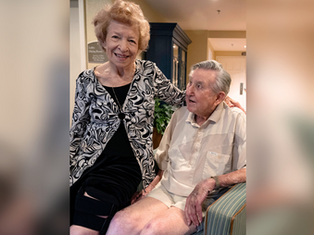 Elderly woman in patterned blouse smiles, stands beside seated man in beige shirt. Indoor setting, warm lighting, friendly atmosphere.