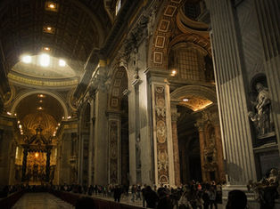 Inside a grand cathedral with ornate columns and arches, people walk beneath intricate ceilings. Warm lighting and statues create a solemn mood.