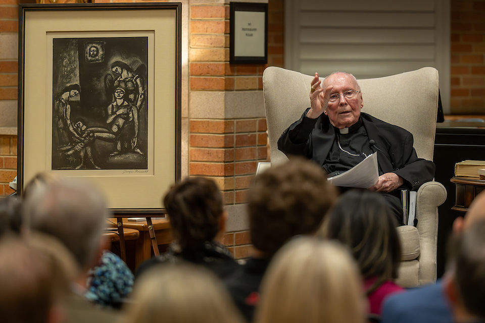 An elderly man in religious attire gestures while speaking in a brick-walled room. A framed artwork is displayed nearby, with an attentive audience.