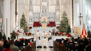 Priests conduct a religious ceremony at a decorated altar with Christmas trees and red poinsettias in a crowded, ornate church.