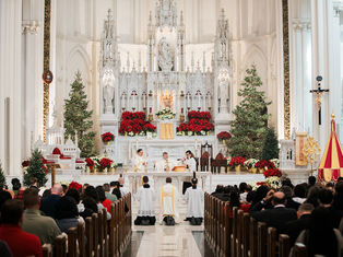 Priests conduct a religious ceremony at a decorated altar with Christmas trees and red poinsettias in a crowded, ornate church.