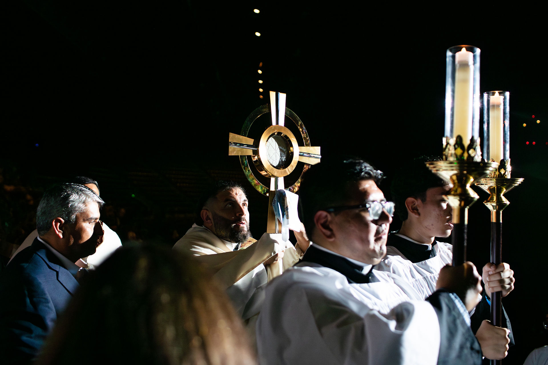 Priests holding a gold monstrance and candles process in a dimly lit setting. The atmosphere is solemn and reverent.
