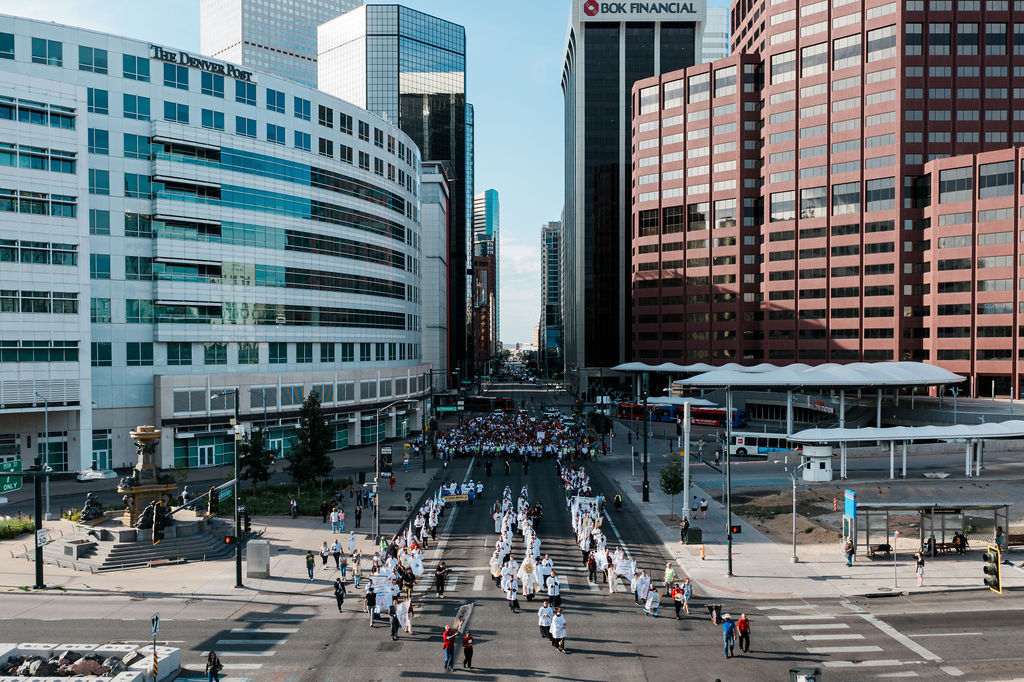 Crowd in white marches down downtown Denver street between The Denver Post and BOK Financial buildings.