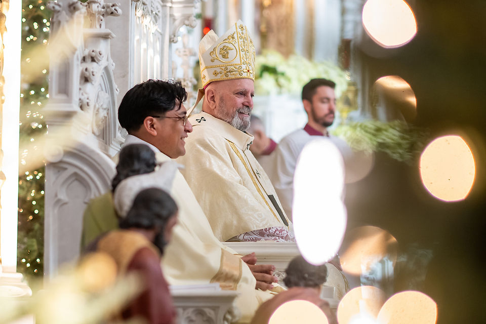 Bishop in ornate robe and hat sits smiling beside others during a church ceremony, with ornate architecture and soft, warm lighting.