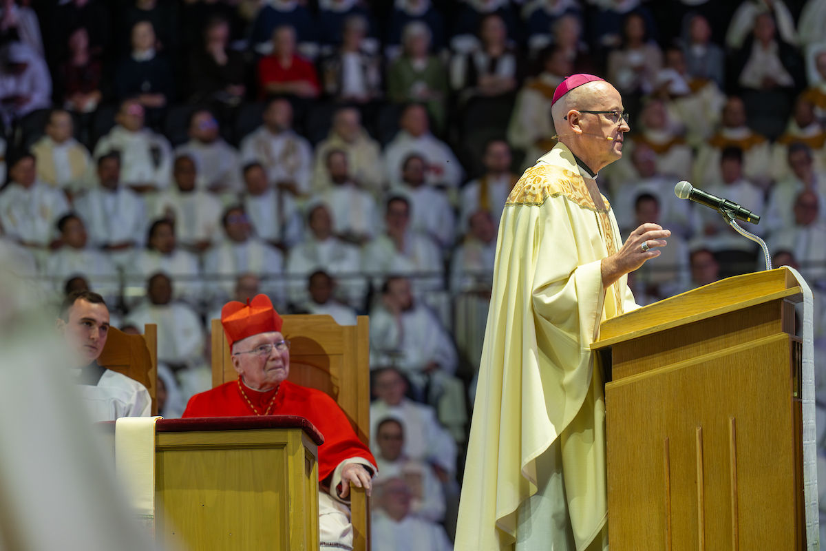Bishop in white and gold robes speaks at a podium during a religious gathering. A cardinal in red sits nearby. Attendees in white fill the background.